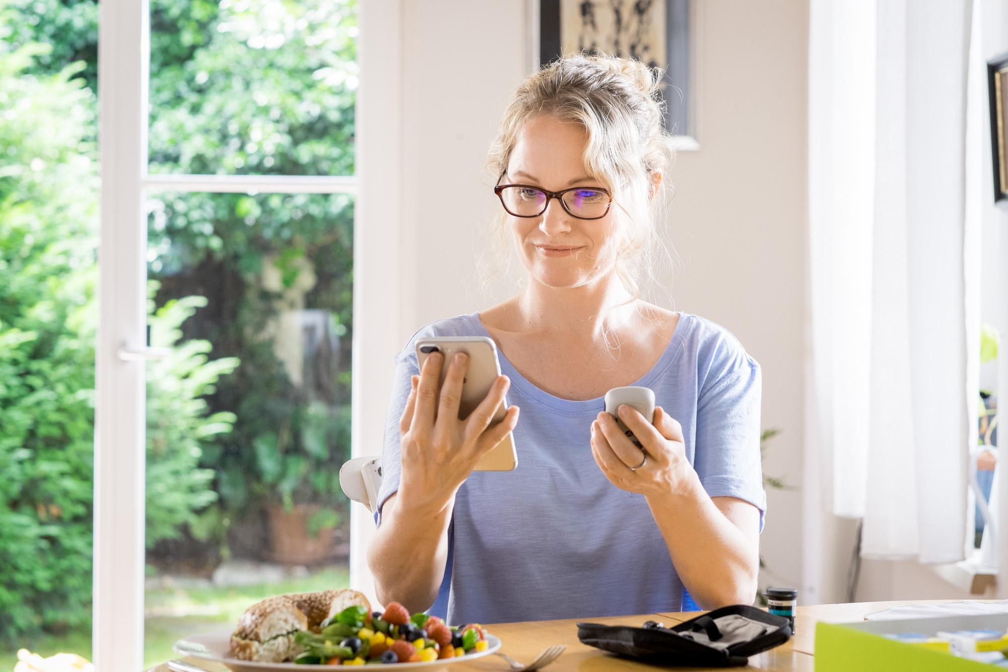 Woman wearing purple shirt and wearing glasses sitting at table next to window. Holding phone and Accu-Chek® Guide meter and there is a plate of food, half a bagel and a salad on the table. There are test strips on the table as well and the corner of the Bundle box.